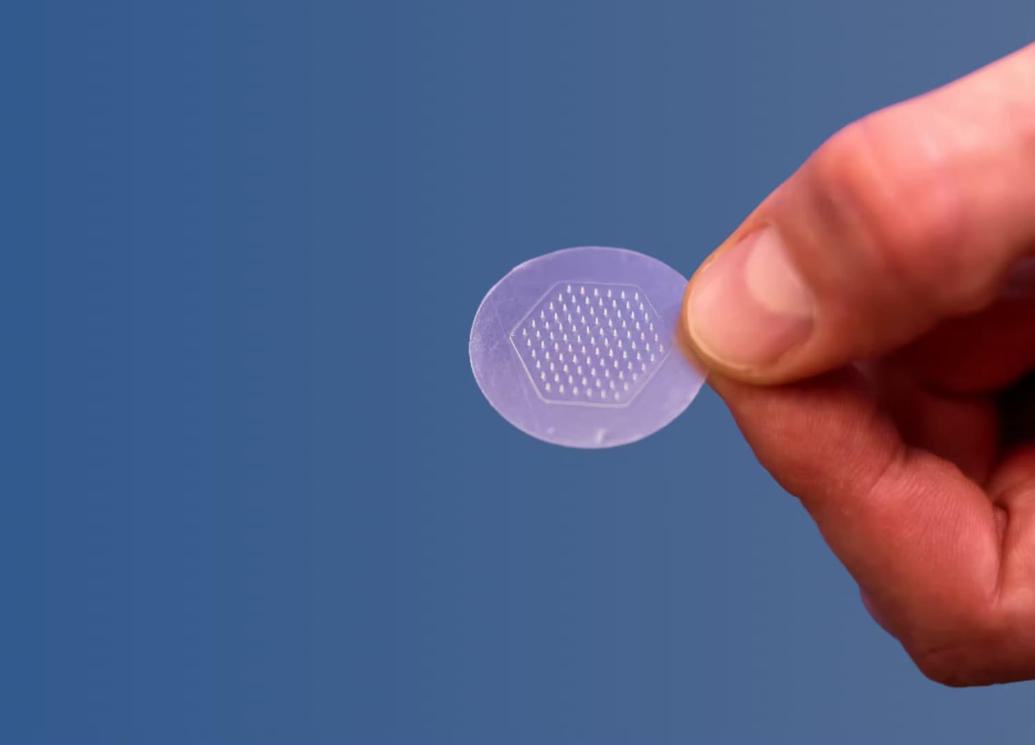 Hand holding a small transparent microneedle patch against a blue background
