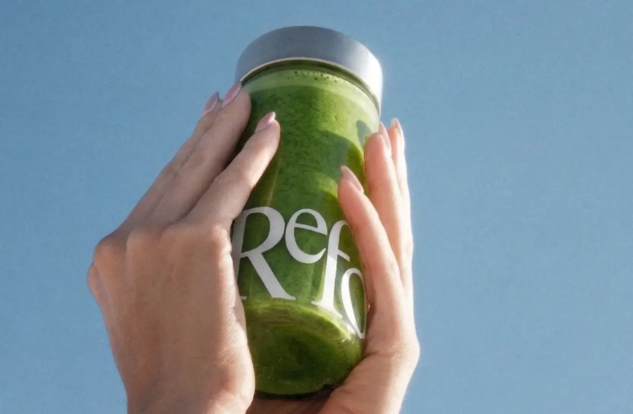 Two hands holding a jar of green Reformed matcha against a blue background